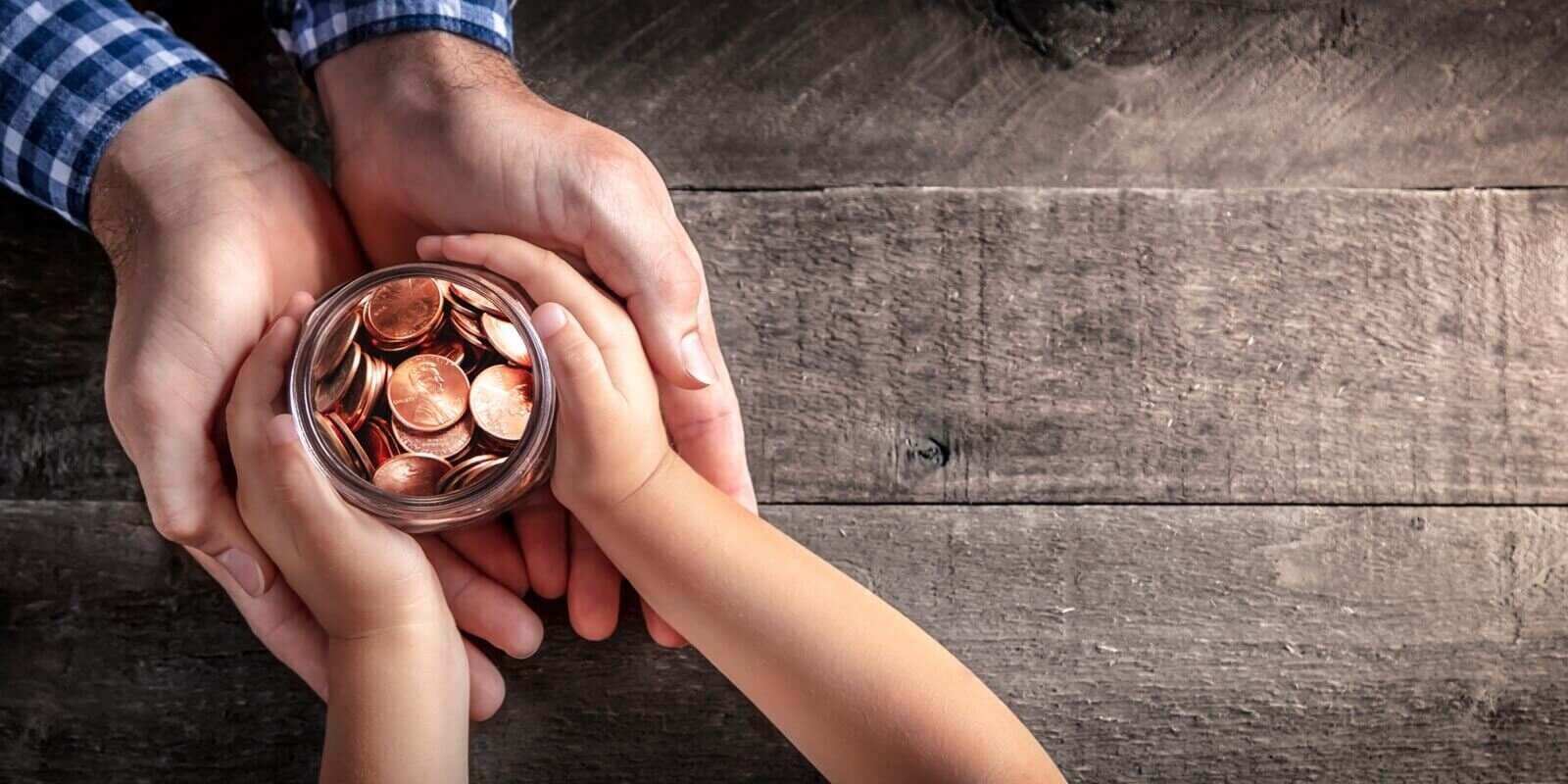 hands of father giving jar of coins to child on wooden table background